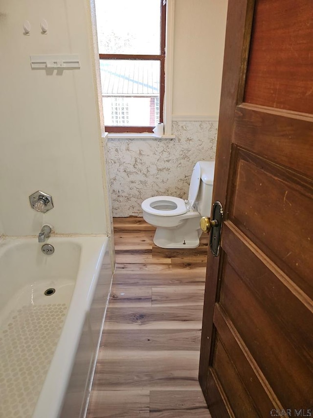 bathroom featuring wallpapered walls, wainscoting, toilet, a bathing tub, and wood finished floors