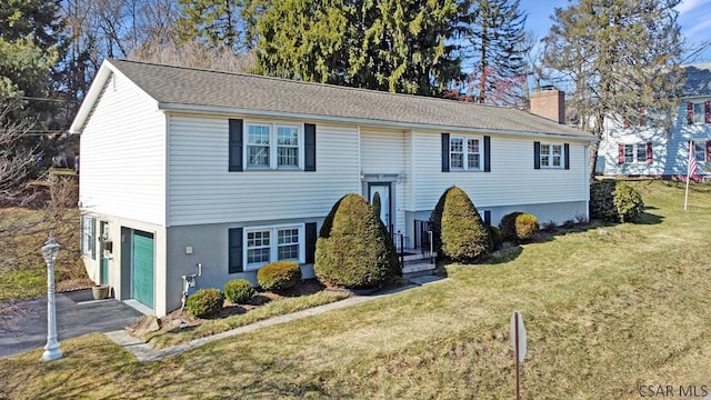 raised ranch featuring a front yard, driveway, an attached garage, a shingled roof, and a chimney