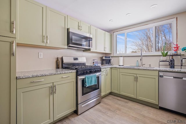 kitchen with stainless steel appliances, a sink, light wood-type flooring, light stone countertops, and green cabinetry