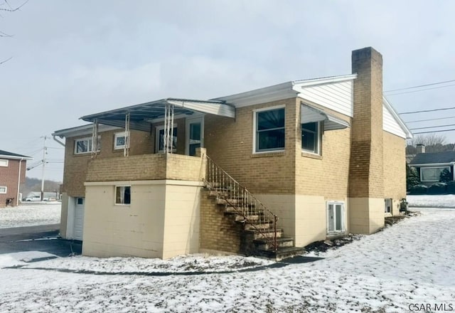 snow covered back of property featuring stairway, brick siding, a chimney, and a garage