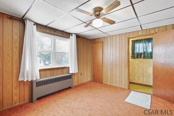 unfurnished room featuring radiator, ceiling fan, wooden walls, carpet floors, and a drop ceiling