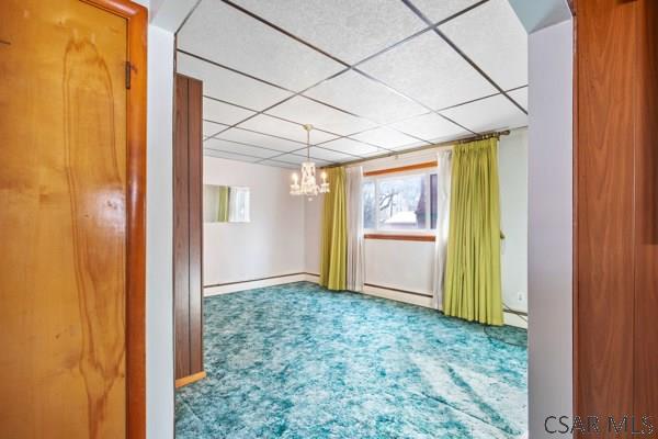 carpeted empty room with a drop ceiling, a baseboard heating unit, and an inviting chandelier