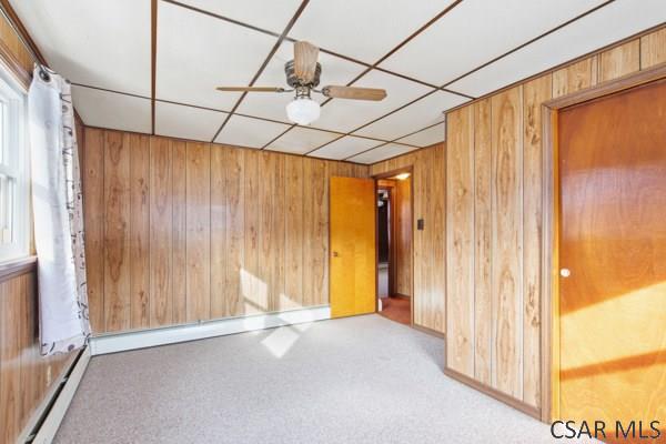 carpeted empty room with a baseboard radiator, ceiling fan, and wooden walls