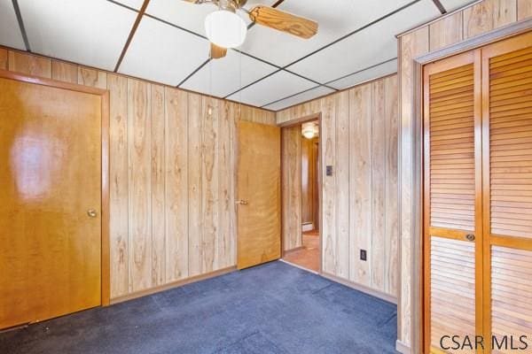 unfurnished bedroom with dark colored carpet, wooden walls, ceiling fan, and a closet