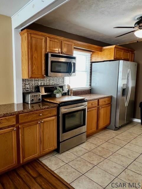 kitchen featuring light tile patterned floors, ceiling fan, appliances with stainless steel finishes, dark stone countertops, and backsplash