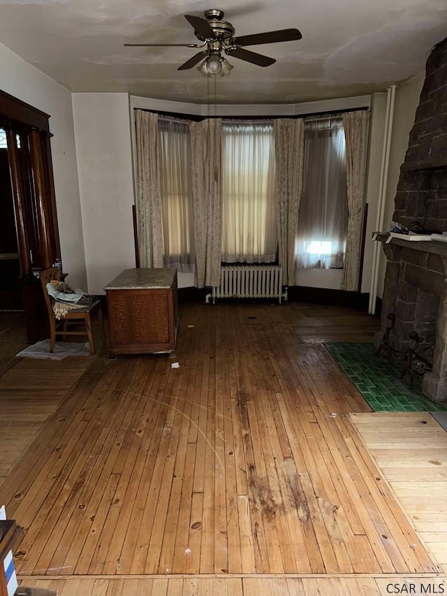 unfurnished living room featuring hardwood / wood-style flooring, a stone fireplace, radiator, and ceiling fan