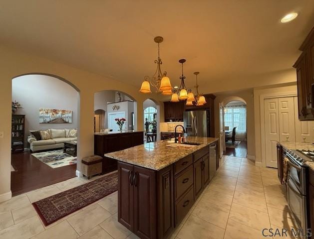 kitchen with appliances with stainless steel finishes, arched walkways, a sink, dark brown cabinets, and a kitchen island with sink