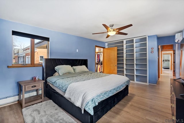 bedroom with wood finished floors, a wall mounted AC, ceiling fan, a spacious closet, and baseboard heating