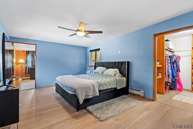 bedroom with a walk in closet, wood finished floors, a closet, a baseboard radiator, and ceiling fan