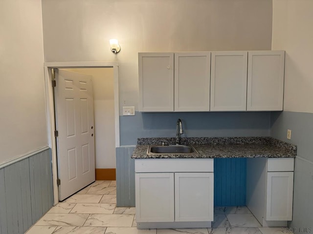 kitchen with marble finish floor, dark stone counters, a sink, and a wainscoted wall