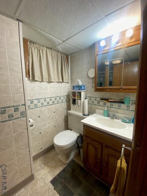 bathroom with a paneled ceiling, tile walls, decorative backsplash, vanity, and toilet