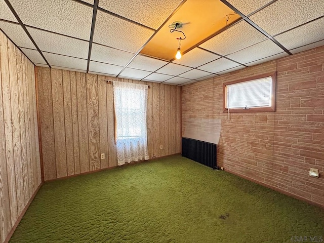 carpeted spare room with a wealth of natural light, radiator heating unit, wood walls, and a drop ceiling