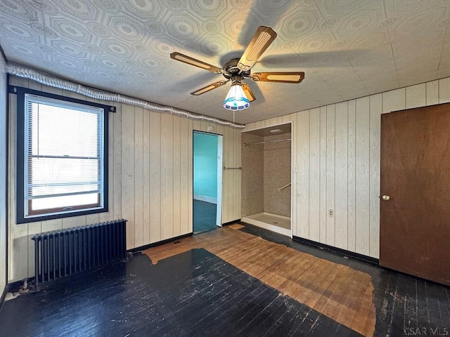 unfurnished room featuring wooden walls, radiator, a ceiling fan, and hardwood / wood-style flooring