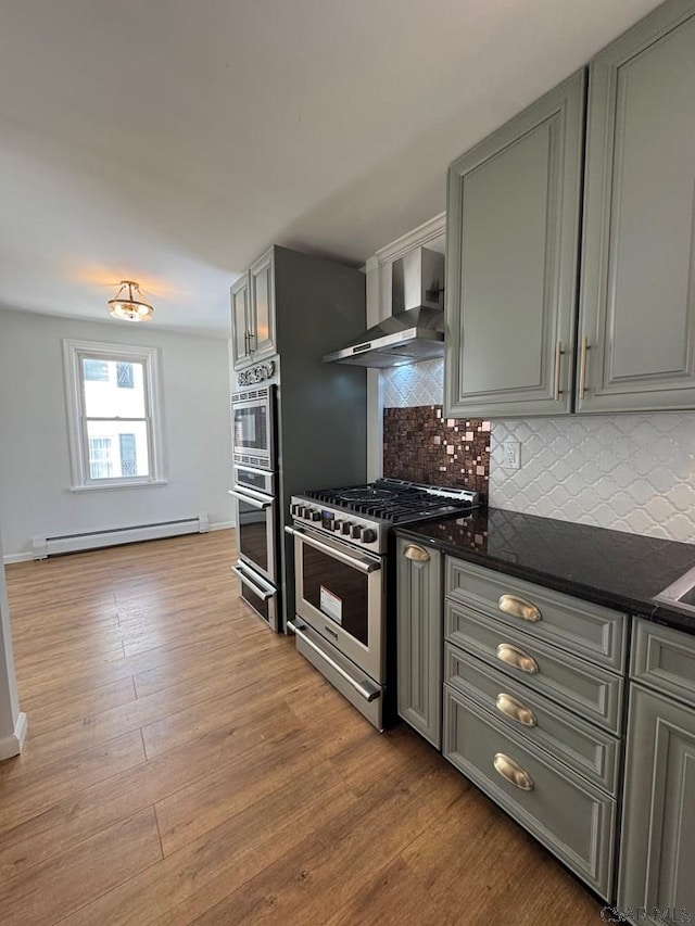 kitchen with appliances with stainless steel finishes, tasteful backsplash, light hardwood / wood-style floors, a baseboard radiator, and wall chimney exhaust hood