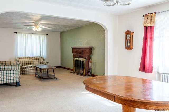 living room featuring baseboards, a fireplace with flush hearth, carpet flooring, arched walkways, and a ceiling fan