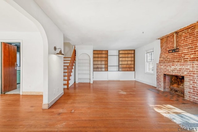 unfurnished living room featuring baseboards, arched walkways, stairway, wood finished floors, and a brick fireplace