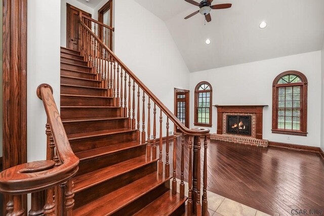 staircase with a brick fireplace, plenty of natural light, high vaulted ceiling, and ceiling fan