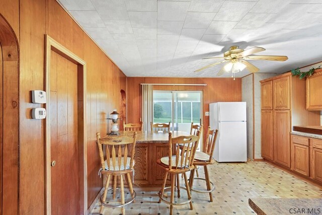 kitchen with a breakfast bar, wooden walls, white refrigerator, ceiling fan, and light stone countertops