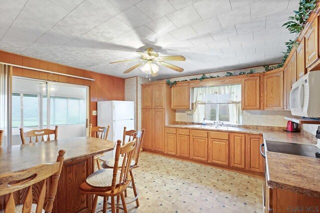 kitchen with wood walls, sink, a kitchen breakfast bar, ceiling fan, and white appliances