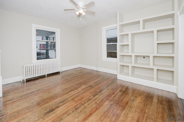 empty room featuring built in shelves, radiator heating unit, ceiling fan, baseboards, and hardwood / wood-style flooring