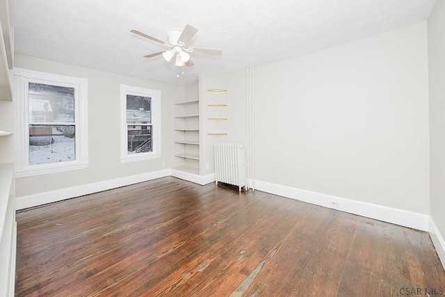 unfurnished room with wood finished floors, a ceiling fan, built in features, baseboards, and radiator heating unit