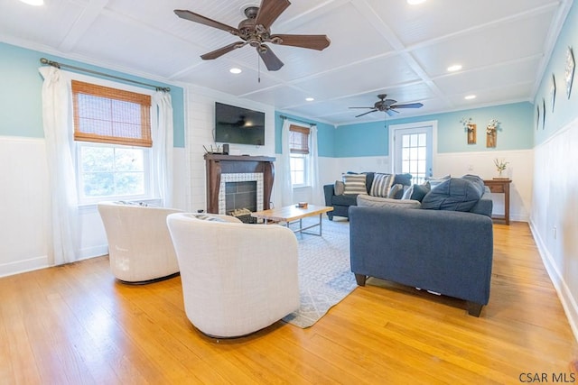 living area with a fireplace, light wood-style floors, ceiling fan, a wainscoted wall, and coffered ceiling