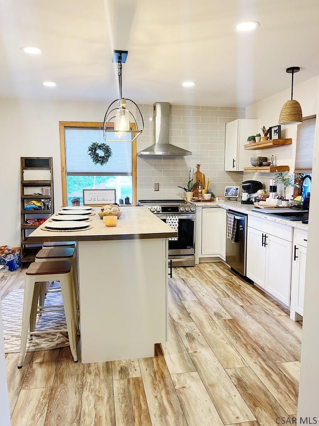 kitchen featuring appliances with stainless steel finishes, a kitchen island, white cabinetry, hanging light fixtures, and wall chimney exhaust hood