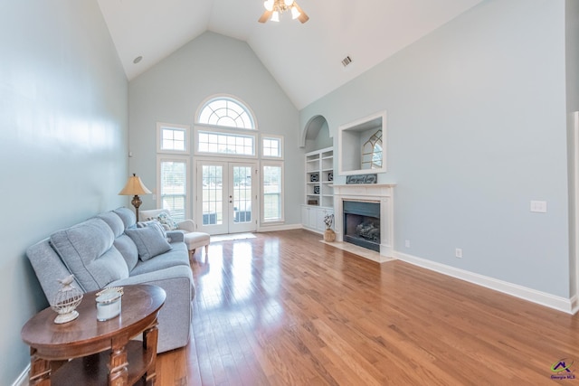 living room featuring ceiling fan, wood-type flooring, a towering ceiling, built in shelves, and french doors