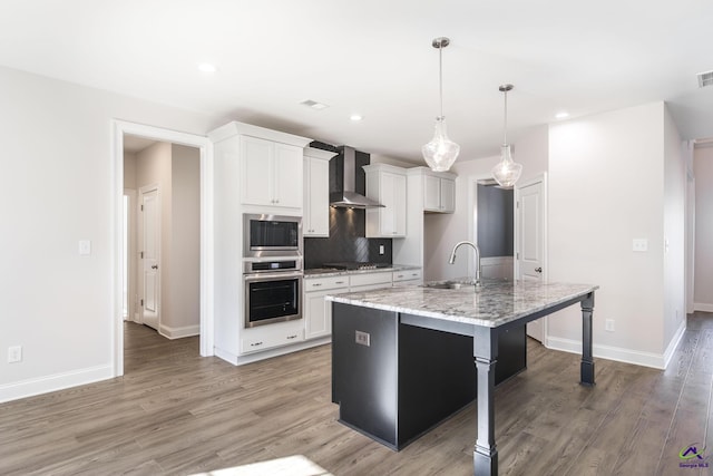 kitchen featuring wall chimney range hood, white cabinetry, appliances with stainless steel finishes, and a kitchen island with sink