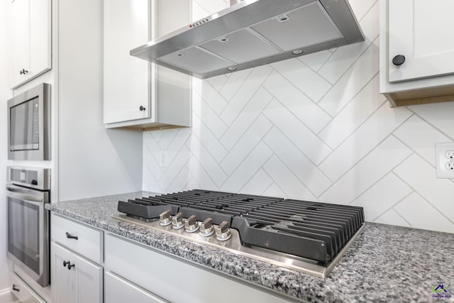 kitchen featuring wall chimney range hood, dark stone countertops, decorative backsplash, white cabinetry, and appliances with stainless steel finishes