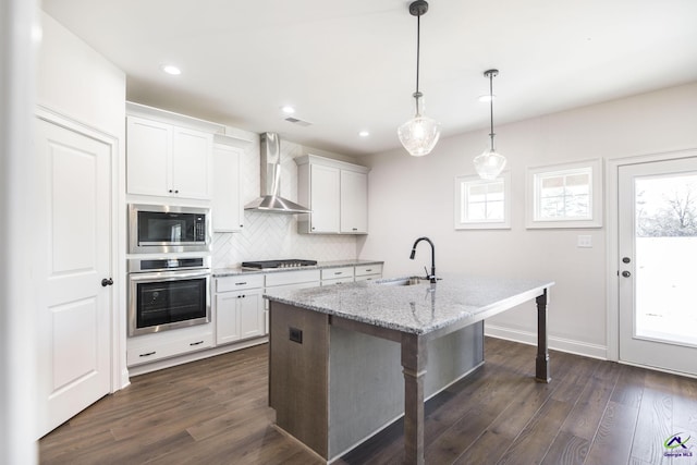 kitchen with an island with sink, stainless steel appliances, wall chimney exhaust hood, white cabinets, and sink