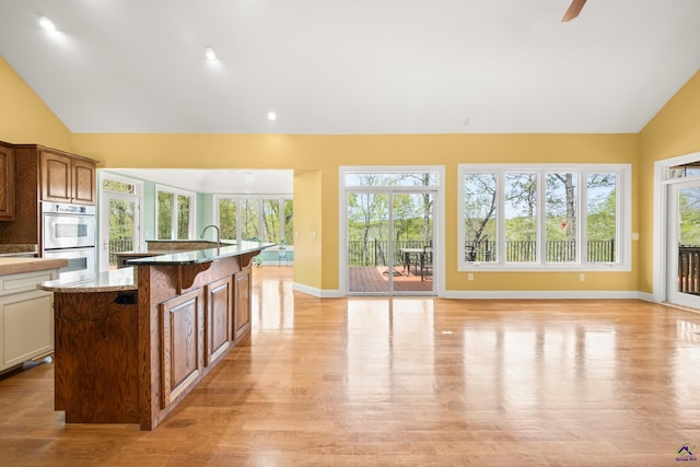 kitchen featuring light stone counters, double oven, vaulted ceiling, a center island with sink, and light hardwood / wood-style floors