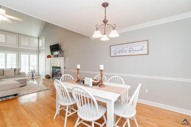 dining space featuring light wood-type flooring, a fireplace, ceiling fan with notable chandelier, crown molding, and vaulted ceiling