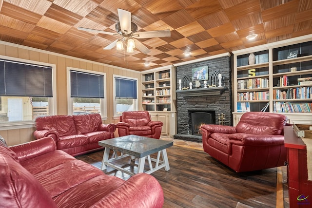 living room with ceiling fan, wooden walls, built in features, dark hardwood / wood-style floors, and a stone fireplace