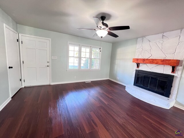 unfurnished living room with ceiling fan, a stone fireplace, and dark wood-type flooring