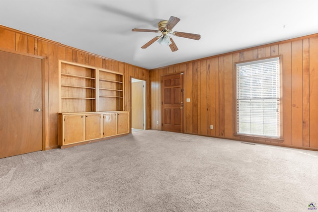 unfurnished living room featuring light colored carpet, built in features, ceiling fan, and wooden walls