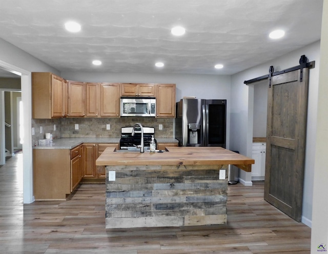 kitchen with light brown cabinets, a barn door, an island with sink, appliances with stainless steel finishes, and butcher block counters