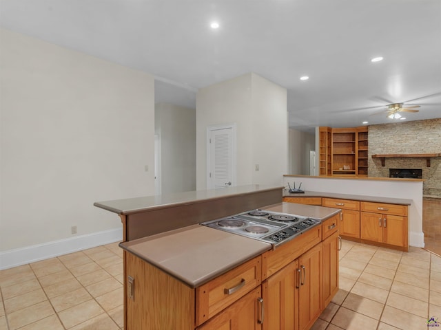 kitchen with stainless steel cooktop, ceiling fan, light tile patterned floors, a center island, and a stone fireplace