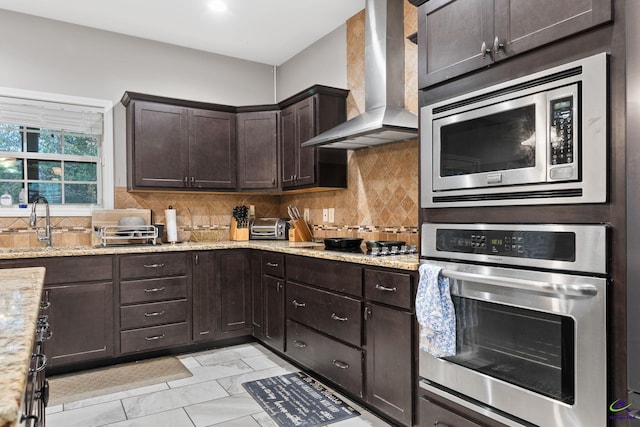 kitchen featuring sink, wall chimney exhaust hood, light stone countertops, appliances with stainless steel finishes, and dark brown cabinetry