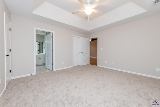 unfurnished bedroom with ceiling fan, ensuite bath, a tray ceiling, light carpet, and a closet