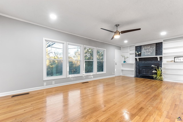 unfurnished living room with ceiling fan, built in features, light wood-type flooring, a brick fireplace, and crown molding