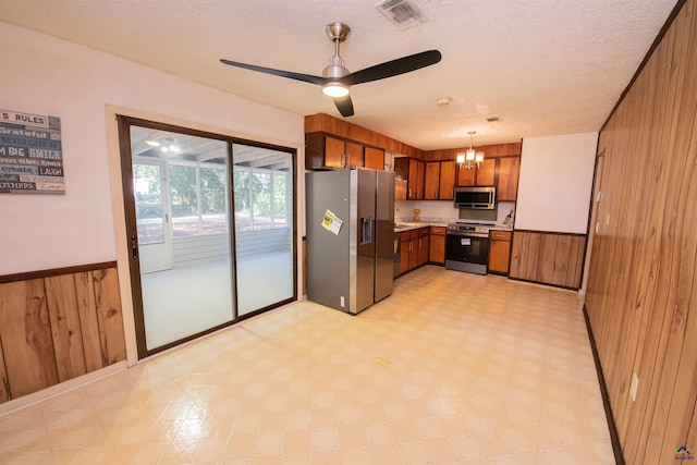 kitchen featuring wood walls, a textured ceiling, pendant lighting, stainless steel appliances, and ceiling fan with notable chandelier