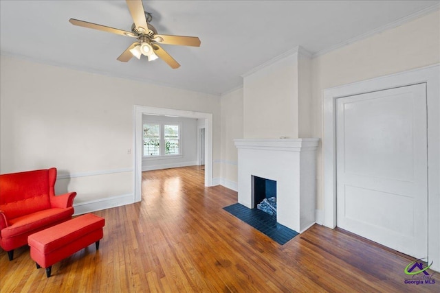 living room featuring baseboards, a ceiling fan, a fireplace with flush hearth, wood finished floors, and crown molding