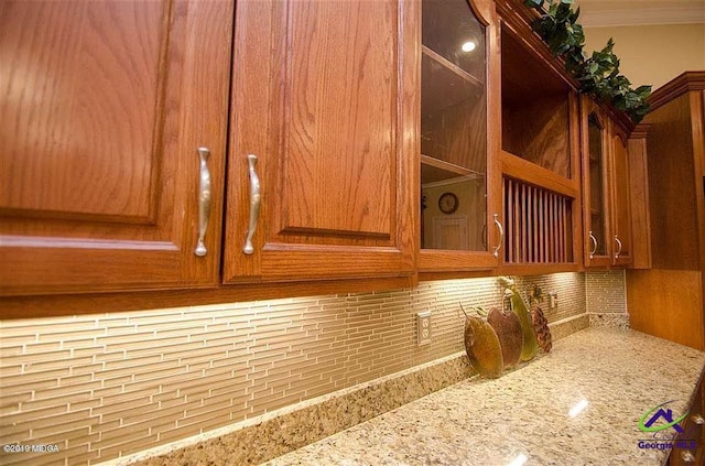 details featuring tasteful backsplash, glass insert cabinets, light stone counters, brown cabinets, and crown molding