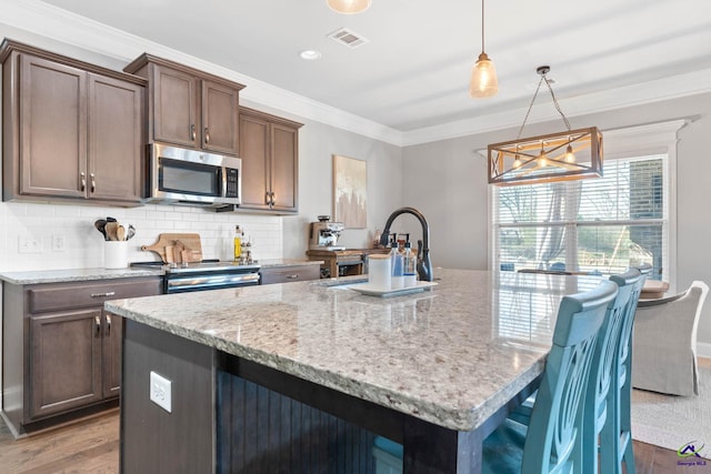 kitchen with decorative backsplash, appliances with stainless steel finishes, ornamental molding, a kitchen island with sink, and wood finished floors