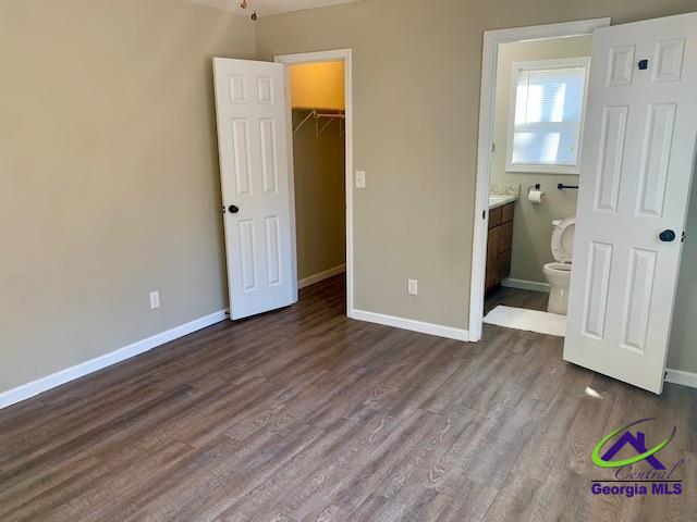 unfurnished bedroom with dark wood-style floors, a closet, a spacious closet, ensuite bath, and baseboards