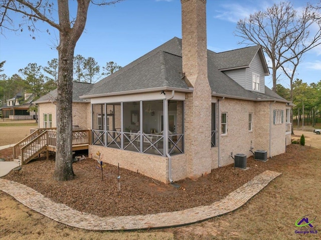 back of property with a sunroom, a chimney, roof with shingles, central air condition unit, and brick siding