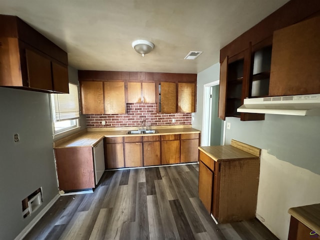 kitchen featuring under cabinet range hood, a sink, backsplash, brown cabinetry, and dark wood finished floors