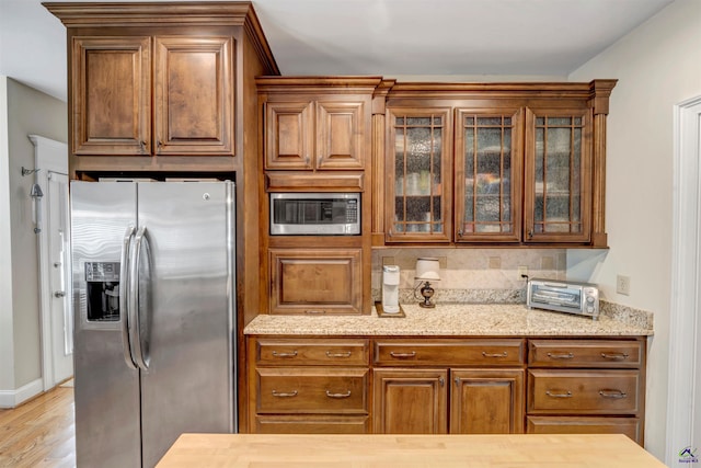 kitchen with brown cabinets, stainless steel appliances, light wood-style flooring, glass insert cabinets, and light stone countertops