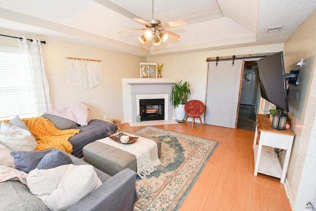 living room with a barn door, a raised ceiling, ceiling fan, a fireplace with flush hearth, and wood finished floors